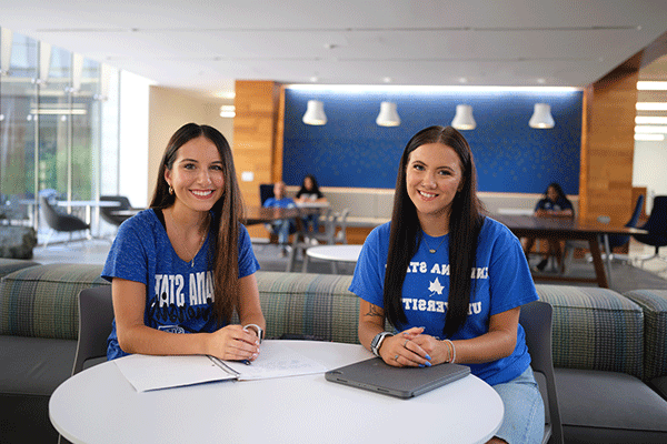 Two white female students wearing blue shirts smile while sitting at a table in the Health and Human Services building.