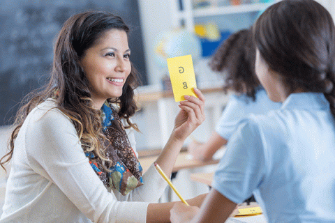 - A smiling woman with dark, medium-length hair and blonde highlights shows a yellow card with the letters ‘G’ and ‘g’ printed on it to a female student seated in front of her. The student has braided black hair and wears a blue shirt. She holds a pencil in her hand. The teacher wears a white long-sleeved top with a multi-colored scarf around her neck. Other students are visible in the background.